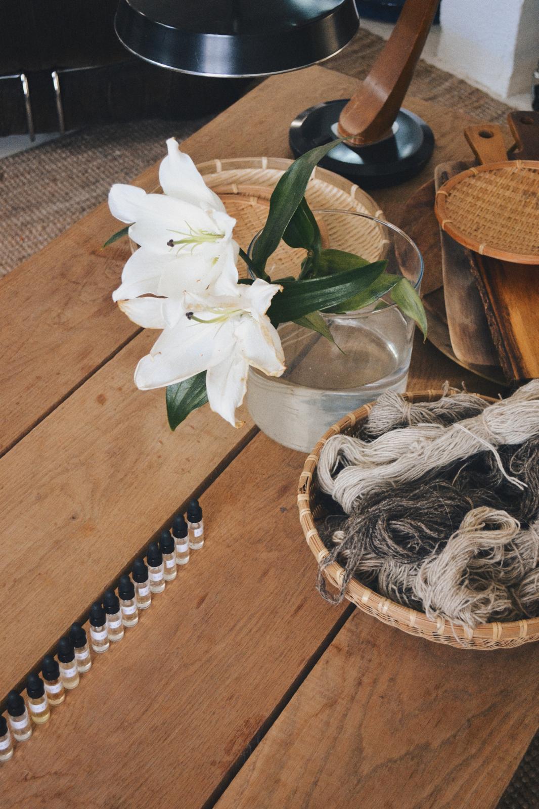 Mesa de madera con dos lirios blancos en un jarrón de cristal, una fila de frascos de muestra con líquido ámbar y una cesta de lana en tonos crudos y grises
